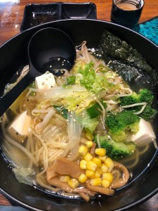 bowl full of ramen broth with noodles, broccoli, sweetcorn, bean sprouts, mushrooms, silken tofu, cabbage and bamboo shoots, with large spoon resting on the side.