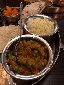 Right hand side of the thali dish. A large metal plate with a large metal bowl in the forground containing a brown curry with peas in. Behind, a bowl of white rice. In the background, a folded chapati, small poppadom, and small bowl with orange curry.