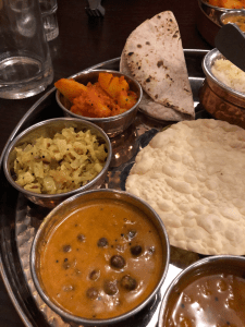 Left hand side of the thali dish. Large metal plate with smaller metal bowls. In the foreground, an orange chickpea curry, on the left, stir-fried cabbage, to the rear, masala potatoes and chapati. on the right, a poppadom.