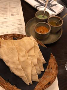 wicker basket with poppadom quarters in, and a small plate with three small bowls containing orange, green, and yellow chutneys.
