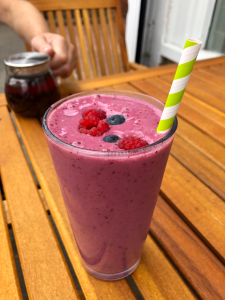 glass filled with bright pink smoothie. Two raspberries and blueberries float on top, with a green and white paper straw.