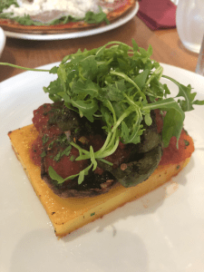 close up of plate with cube of cooked polenta on, topped with vegetables in tomato sauce and green salad leaves.