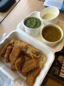 White takeaway container opened up. On the left side are six vegan chicken nuggets and a battered sausage. On the right side are three smaller, round containers filled with three different sauces.