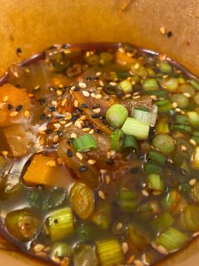 cardboard pot filled with dark brown broth, vegetable and cabbage chunks, sliced spring onion and topped with black and white sesame seeds