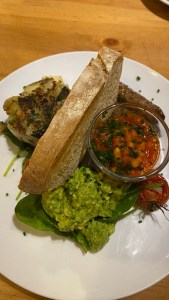 Plate with toast standing upright in centre. on the left, ball of fried mashed potato. On the right, a small glass dish with baked beans in topped with chives; one roasted vine tomato, one sausage visible and pile of smashed avocado on spinach leaves.