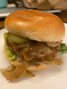 shiny brioche bun sandwiching a messy looking burger. Lettuce leaf on the bottom, with burger patty and pile of onions and sauce spilling out, with gherkin slice on top.