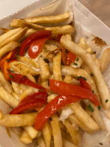 box filled with thin cut fries, topped with slices of red pepper, some hints of cheese sauce and sprinkling of chives
