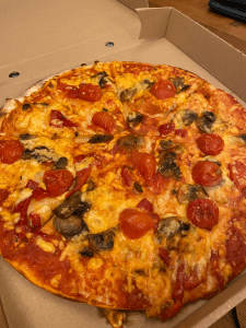 Open pizza box with pizza covered in yellow-orange melted looking cheese, cooked cherry tomato halves and chunks of brown mushrooms