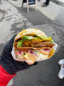 gloved hand holding up english muffin stuffed full of avocado slices, brown scrambled tofu, chinks of raw onion, two tortilla chips, and a pale pink sauce.