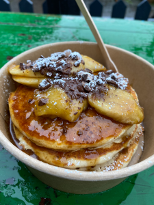 round cardboard bowl with wooden fork sticking out of it, stuck into stack of three large pancakes, shiny from caramel sauce topping. Slices of banana with dark chocolate grated on top.