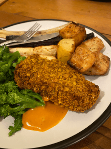 plate with roast potatoes, parsnips, purple sprouting broccoli, and a crispy chick*n fillet on. Some cajun mayo next to the fillet and a knife and fork resting on the side.