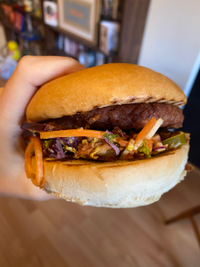 hand holding up a burger in a thick brioche style bun, beef style patty on top of thick bed of shredded vegetable coleslaw (carrots, red cabbage, pickles visible)