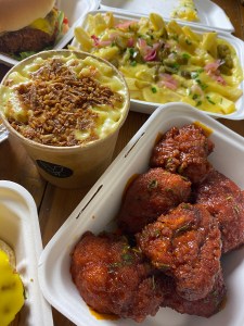 cauliflower balls in a red breadcrumbed coating, top of a cardboard pot full to the very top with yellow pasta and topped with brown crispy onions, white takeaway pot with chips coated in cheese sauce, green jalapenos and pink pickled onions.