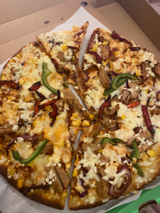 pizza in a box, burnt round the edges, covered in grated cheese, red onion, sweetcorn kernels, mock chicken and burnt red and green bell pepper slices.