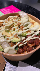 Round cardboard bowl full to the brim with vegetables. Visible on top are dark brown crispy seasoned chickpeas, slices of pale green avocado, two pickles. Smothered in light green mayo dressing.